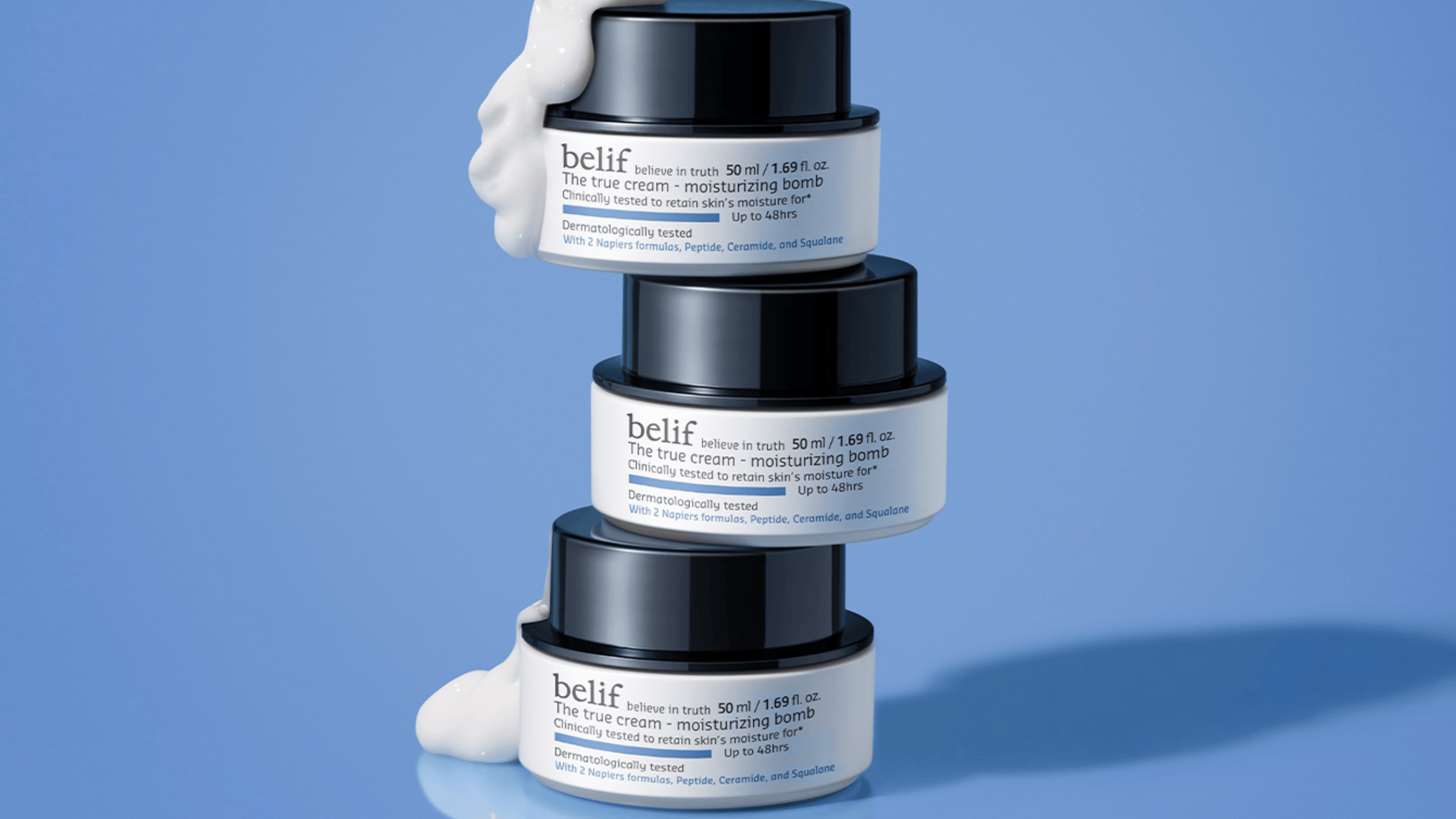 Tre staplade burkar av "belif The True Cream - Moisturizing Bomb" på blå bakgrund. Översta burken har kräm som rinner över kanten, vilket visar dess återfuktande egenskaper. Produkten är testad för att bibehålla hudens fukt i upp till 48 timmar.