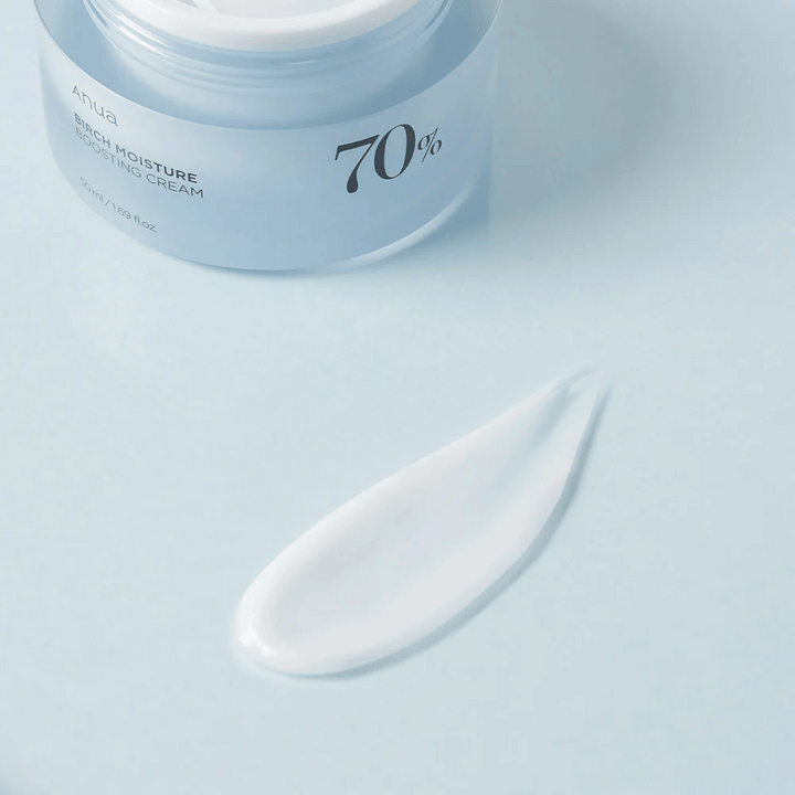 Bilden visar en burk av "Anua Birch Moisture Boosting Cream" som innehåller 70% av den aktiva ingrediensen. Burken är öppen och bredvid den finns en swatch av krämen på en yta, vilket ger en indikation på produktens konsistens och färg. Designen är ren med en mjuk blå toning som matchar namnet som antyder fuktgivande egenskaper från björk.