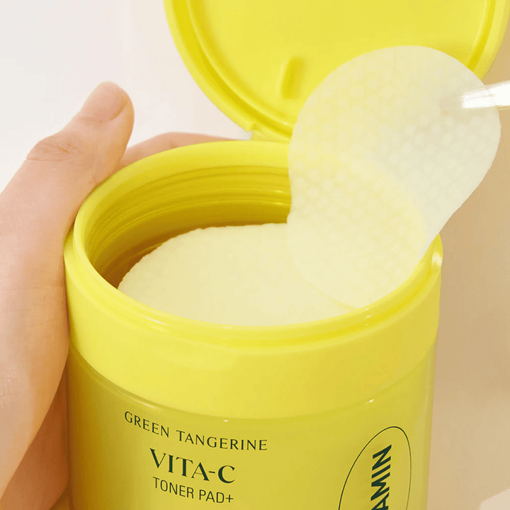 En hand använder en plastpincett för att plocka upp en tonerpad med textur från en ljusgul behållare märkt "GREEN TANGERINE VITA-C TONER PAD+" av märket "gooda