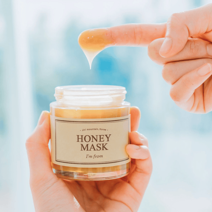 En hand håller öppen burk av "I'm from Honey Mask", och en finger dypps i den gyllene masken mot en ljus bakgrund.