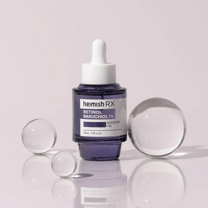 Heimish RX Retinol Bakuchiol Booster Oil 1% i en elegant lila glasflaska med pipett. Oljan är placerad på en reflekterande yta med glaskulor runtom, vilket ger en lyxig och modern känsla.